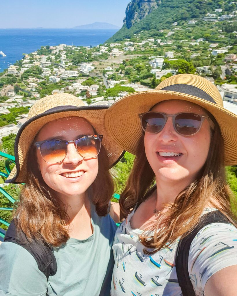 Krissie and Katherine smiling with sun hats and sunglasses standing in front of a view over Capri, Italy. The blue ocean can be seen in the distance with a big cliff on the right side and lots of buildings and green plants covering the landscape "See Naples On a Budget With These Six Great Ideas!"