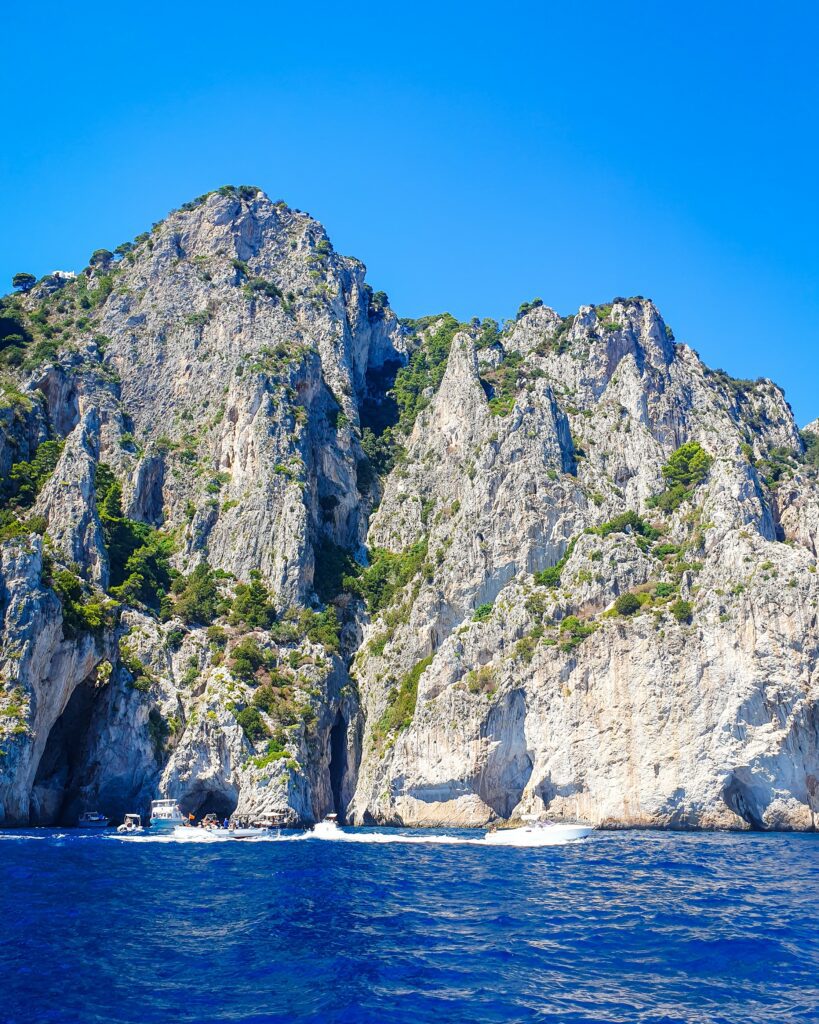 Some big white cliffs with green plants on top behind the beautiful blue ocean in Capri, Italy "See Naples On a Budget With These Six Great Ideas!"