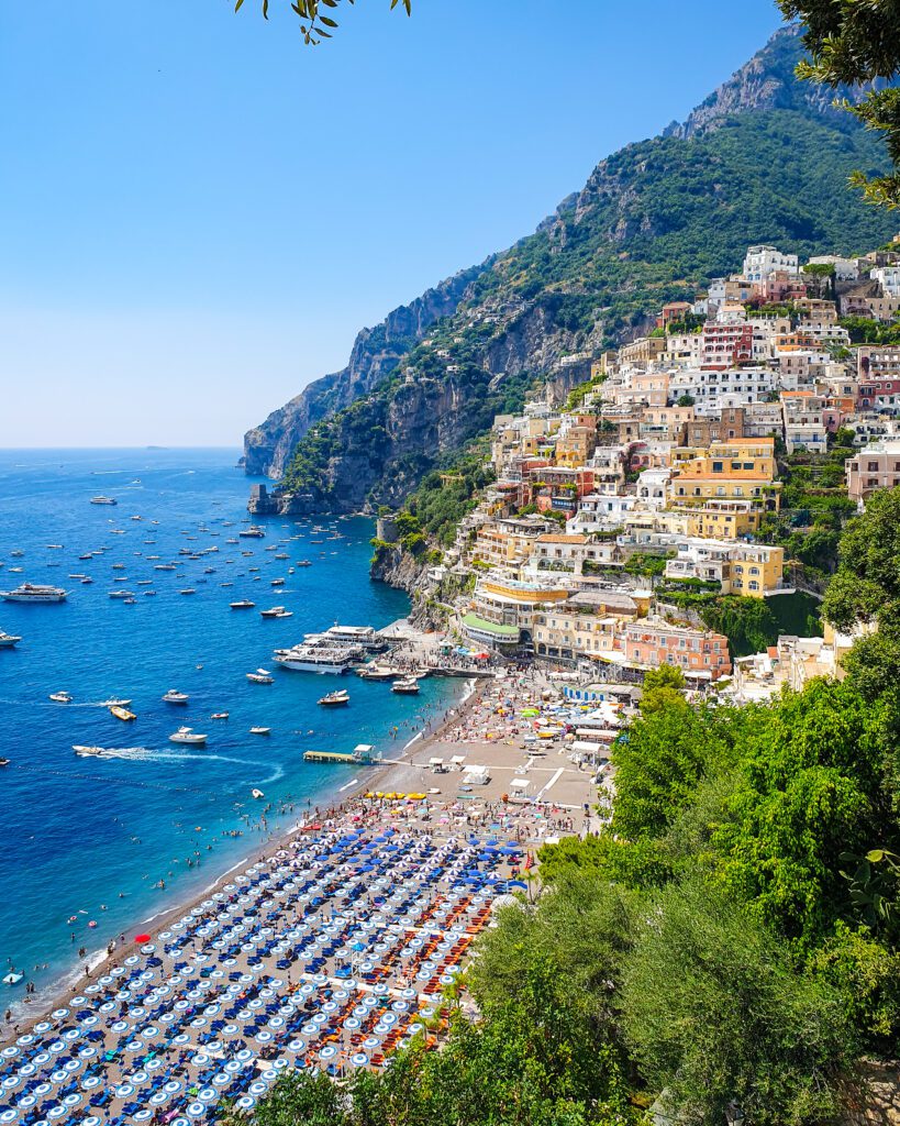 A view over Positano on the Amalfi Coast, Italy. There is a big beach with hundreds of umbrellas, a beautiful blue ocean with heaps of boats on it and a big cliff half taken up with lots of colourful buildings "See Naples On a Budget With These Six Great Ideas!"