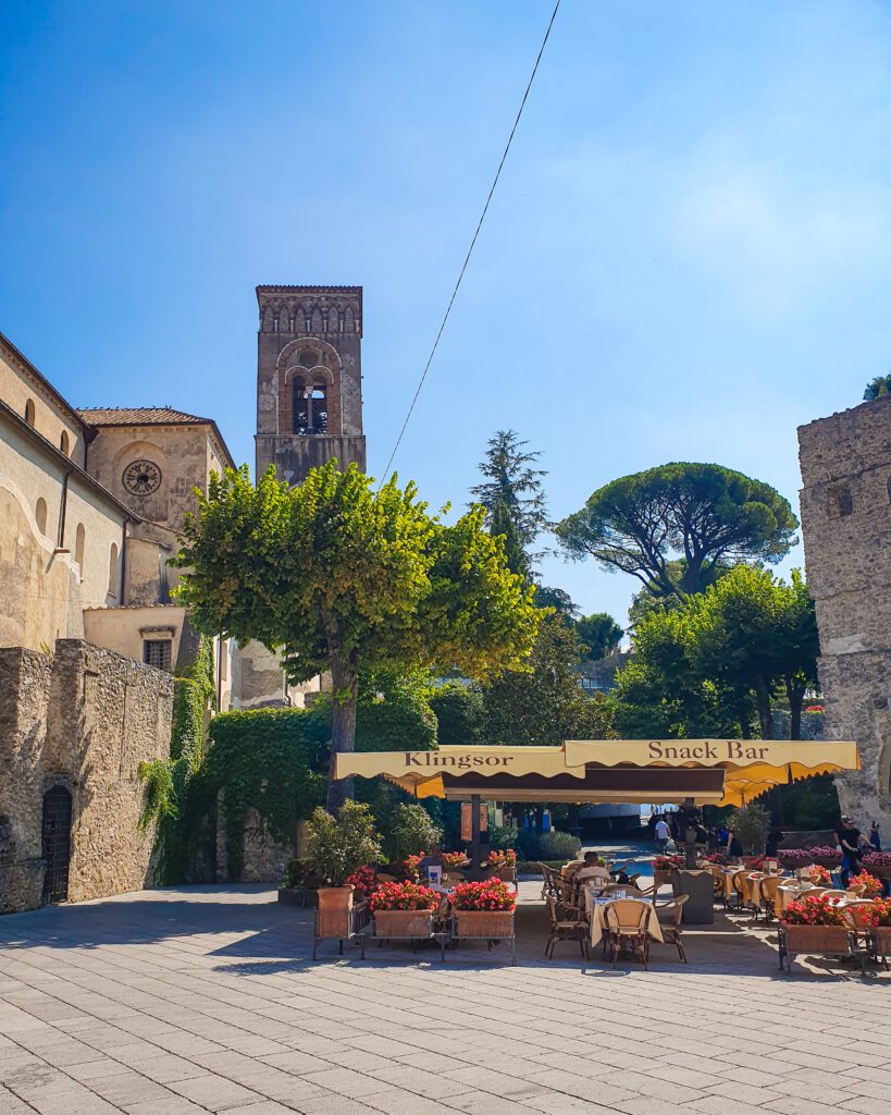 A picture of a little town square in Ravello on the Amalfi Coast, Italy. In the picture is an outdoor seating area of a restaurant, some trees, and some stone buildings including a bell tower "See Naples On a Budget With These Six Great Ideas!"