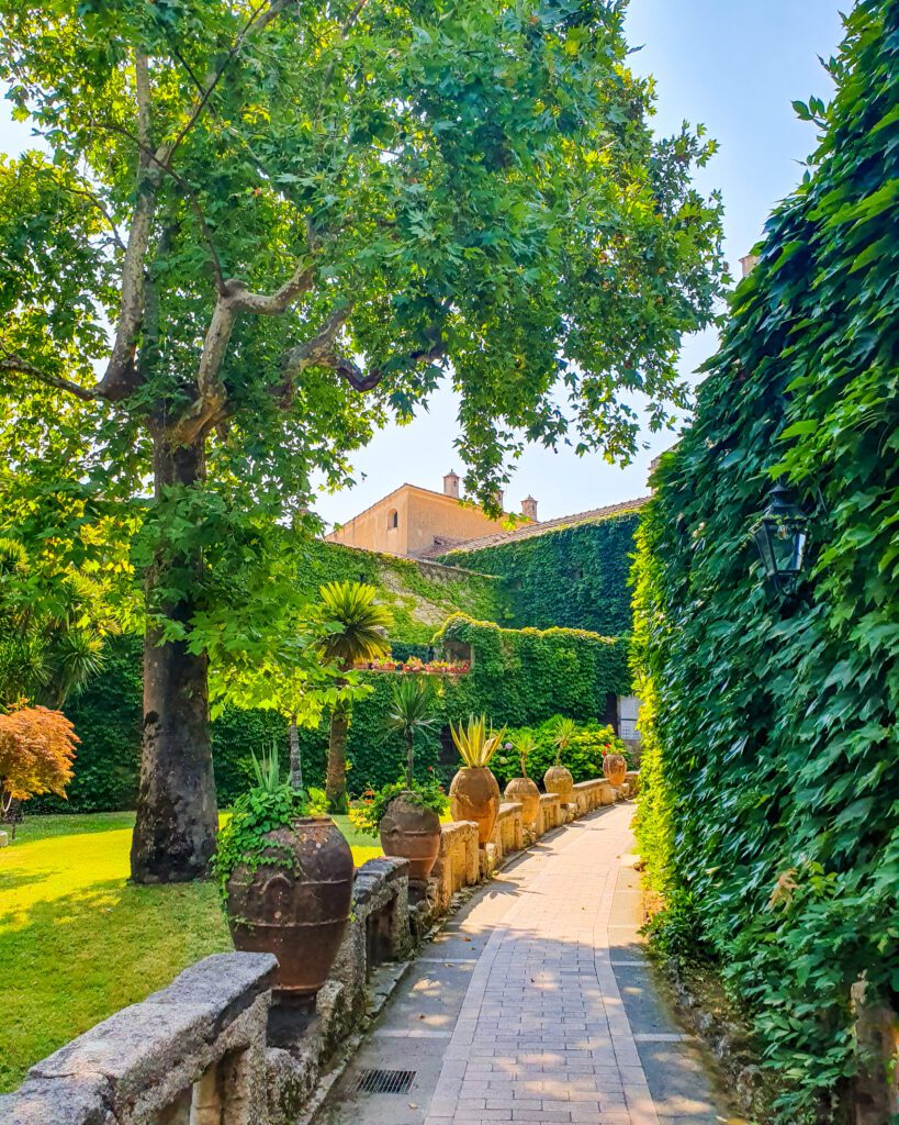 A picture of a lush green garden with trees, a patch of grass on the left, lots of potted plants, a path leading through it and a tall green bush on the right at Villa Cimbrone, Ravello which is on the Amalfi Coast, Italy "See Naples On a Budget With These Six Great Ideas!"