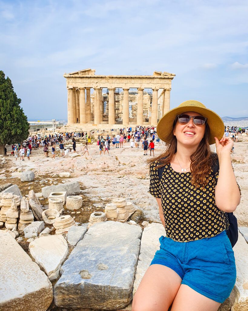 Katherine sitting on a stone wall and smiling in Athens, Greece. Behind her is the Parthenon and lots of people walking around it "How to Make the Most of the Athens Combined Ticket"