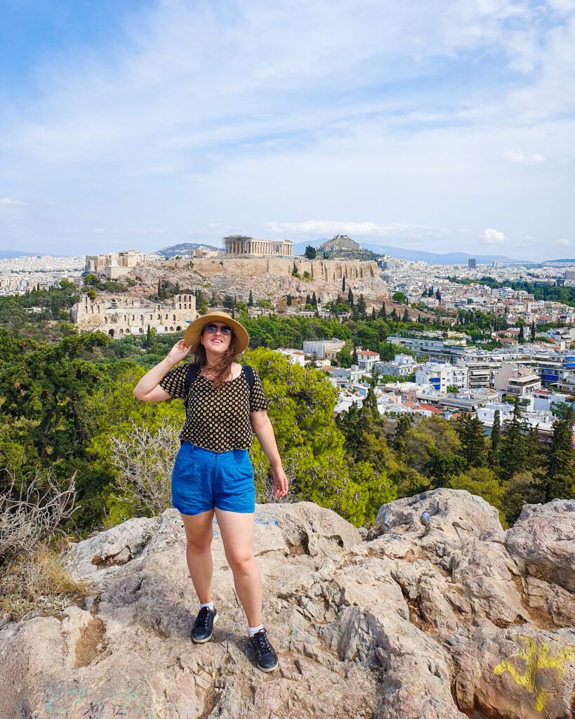 Katherine smiling and standing on a rock on a hill with trees and white buildings behind her. Beyond those is the Acropolis hill with the Parthenon on top in Athens, Greece "How to Make the Most of the Athens Combined Ticket"