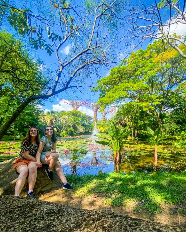 Katherine and Krissie sitting on a rock in front of the Gardens By The Bay super trees in Singapore. In front of the super trees is a pond with lots of lily pads on it and lots of trees and grass around "Singapore: A Complete Guide to the Public Transport System"