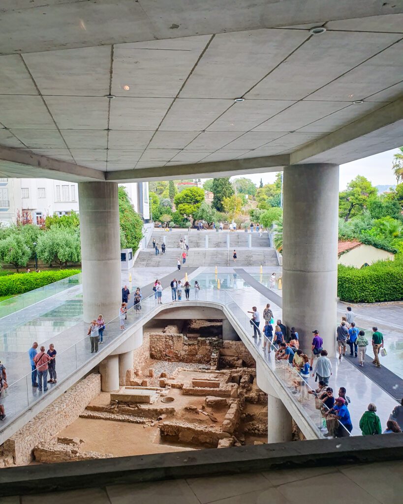 A view of the outside of the Acropolis museum which has 2 huge columns and a glass barrier in an arched shaped between them. Below the glass barrier you can see the old stone ruins of an ancient neighbourhood in Athens, Greece "The 6+ Best Budget Friendly Activities in Athens"