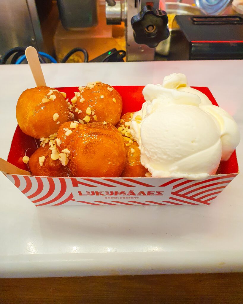 A tub of golden brown doughnut balls with a scoop of vanilla ice cream on the side and crushed nuts on top. The cardboard container they're in says Lukumades on it which is a great cafe in Athens, Greece "The 6+ Best Budget Friendly Activities in Athens"