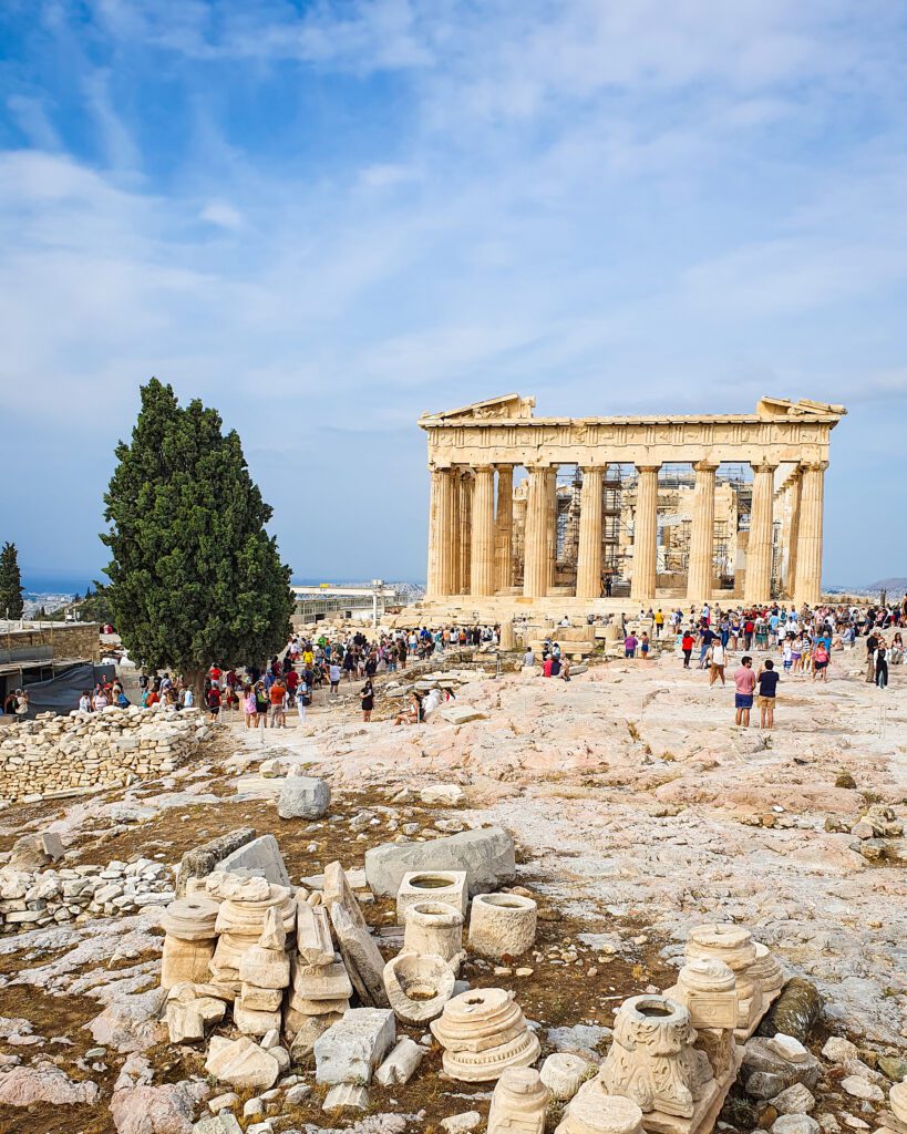 A view of the Parthenon with lots of rocks and people around and a big green tree on the left of the photo in Athens, Greece "The 6+ Best Budget Friendly Activities in Athens"