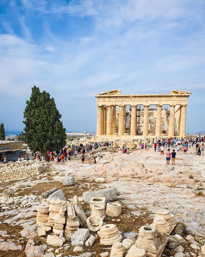 A view of the Parthenon with lots of rocks and people around and a big green tree on the left of the photo in Athens, Greece "The 6+ Best Budget Friendly Activities in Athens"