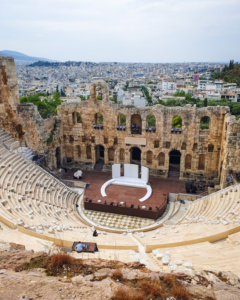 A sloped amphitheatre in Athens, Greece. There is bench seating for heaps of people with a big backdrop made up of lots of stone archways. They're setting up a stage at the bottom of the sloped seating and behind the amphitheatre is a view over Athens which is a sprawling city with lots of white buildings ""How to Make the Most of the Athens Combined Ticket""