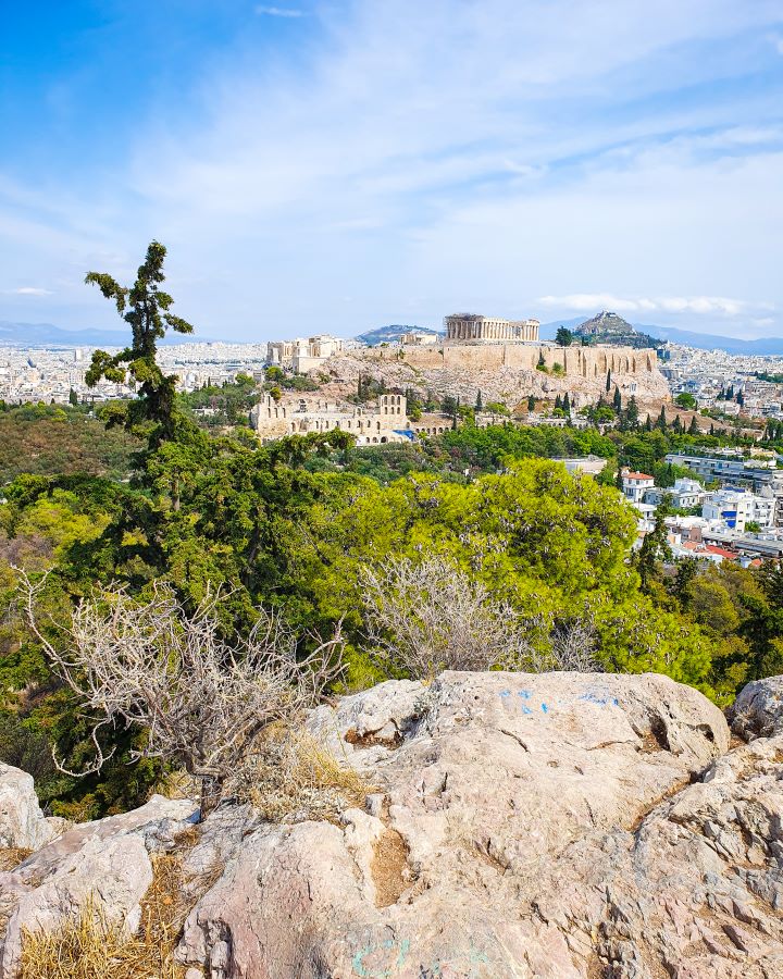 A view from Philoppapos Hill where you can see the Acropolis hill with the Parthenon on top. In front of the Acropolis are lots of green trees and you can see the white buildings of Athens, Greece all around "The 6+ Best Budget Friendly Activities in Athens"