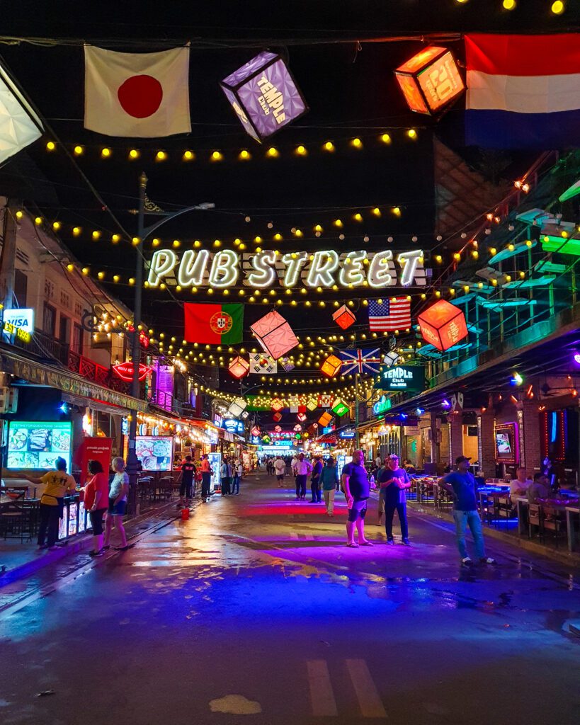 A view of Pub Street in Siem Reap, Cambodia that is taken at night. There is a huge light up sign saying Pub Street over the street and lots of bright coloured lights lit up with people walking underneath and sitting at the restaurants along the street "The Ultimate Guide to Siem Reap: Temples, Markets, and More!"