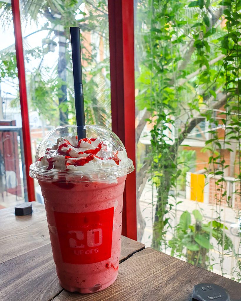 A picture of a pink frappe with cream and a red sauce drizzled on top. The frappe is in a plastic takeaway cup with a straw and behind it is some green vines on the outside of Temple Coffee n Bakery in Siem Reap Cambodia "The Ultimate Guide to Siem Reap: Temples, Markets, and More!"