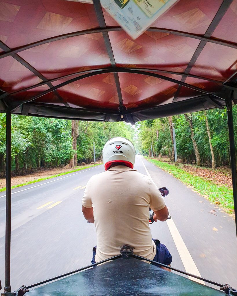 A picture taken while Katherine and Krissie were on the back of a tuk tuk in Siem Reap Cambodia. You can see the roof of the tuk tuk with the driver in a helmet in the middle of the photo. The tuk tuk is on an empty road with lots of bushy trees around "The Ultimate Guide to Siem Reap: Temples, Markets, and More!"