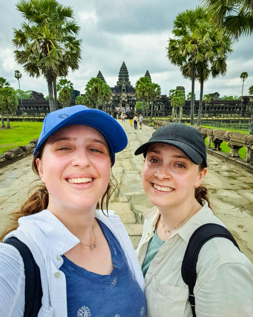 Katherine and Krissie standing in front of the Angkor Wat temple in Siem Reap, Cambodia with lots of trees and grass as well. They're both wearing caps and looking very hot and sweaty! "The Ultimate Guide to Siem Reap: Temples, Markets, and More!"