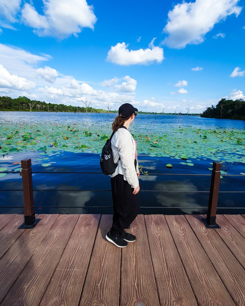 Krissie standing on a wooden walkway with a huge lake filled with lily pads behind her. The walkway goes from the land over to the Neak Pean temple in Siem Reap Cambodia "The Ultimate Guide to Siem Reap: Temples, Markets, and More!"