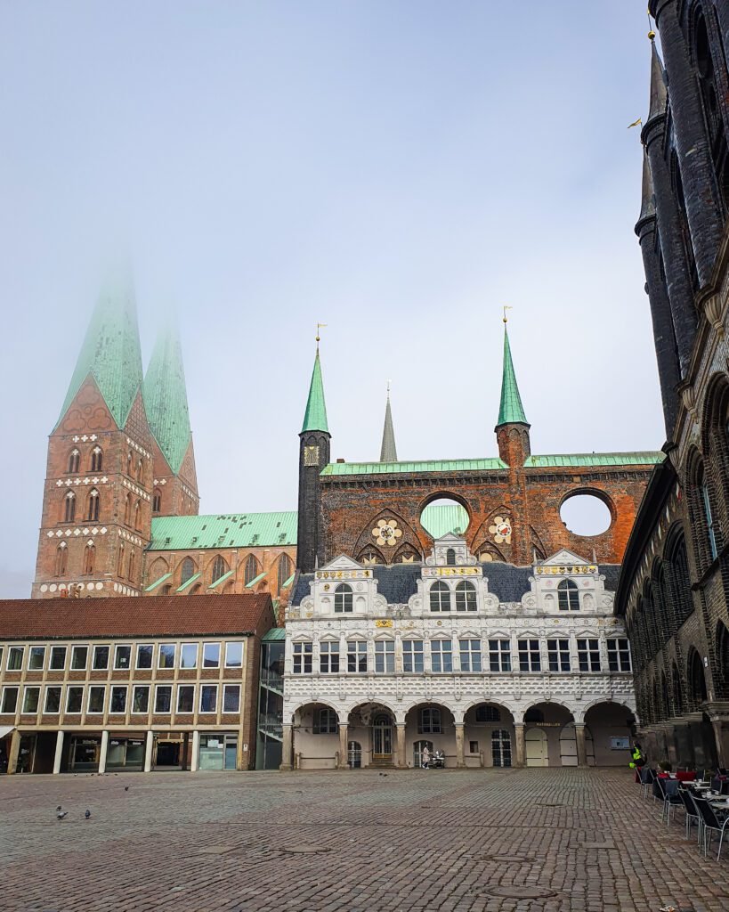 St Mary's Cathedral in Lübeck, Germany with its green spires and red brick building which can be seen across a large cobbled town square "How to See Lübeck in a Day Trip from Hamburg"