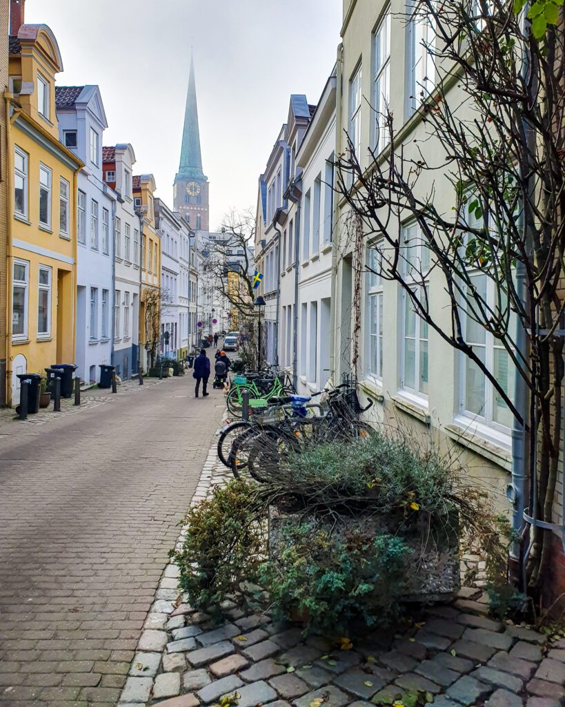 A street in Lübeck with some colourful old European buildings, some plants and a church spire in the background "How to See Lübeck in a Day Trip from Hamburg"