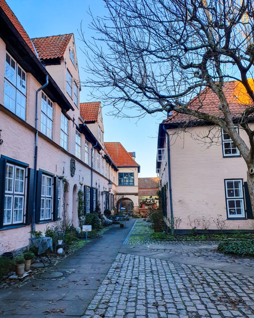 The Füchtingshof courtyard in Lübeck, Germany. It has a cobbled street and pink buildings around it with orange tiled roofs "How to See Lübeck in a Day Trip from Hamburg"