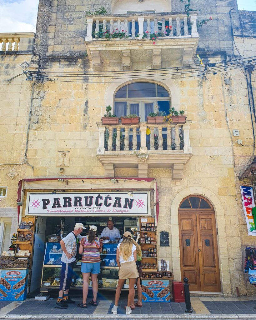 Some people standing at a Maltese bakery in Rabat, Malta with a sign above saying "Parruccan" "Eight Amazing Things to See in Malta"