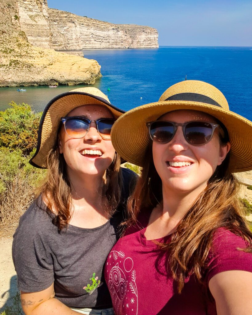 Krissie and Katherine smiling with sunglasses and big hats standing in front of a beautiful view with the blue ocean and a big white cliff behind them in Gozo, Malta "Eight Amazing Things to See in Malta"