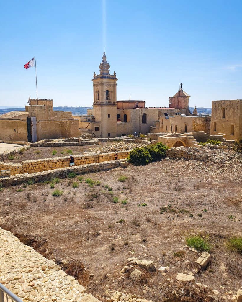 The sandstone coloured Cathedral of the Assumption in Victoria in Gozo, Malta with the Maltese flag flying over the building "Eight Amazing Things to See in Malta"