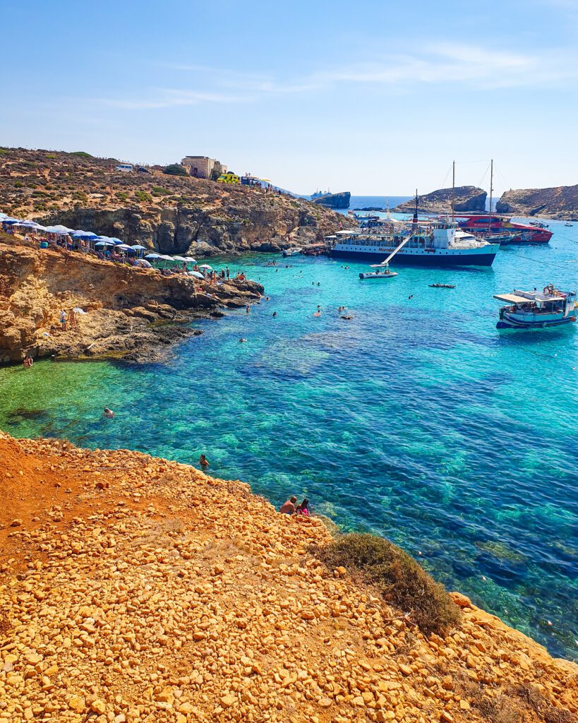 The Blue Lagoon on the island of Comino in Malta. There's boats and people in the water and the water is a beautiful clear blue and green colour with orange rocks around it "Eight Amazing Things to See in Malta"