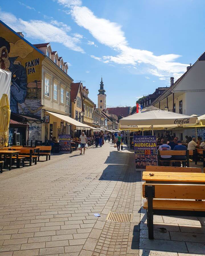 A street in Zagreb, Croatia with lots of restaurants and outdoor seating with people sitting and enjoying the blue sky "How to Affordably See Zagreb"