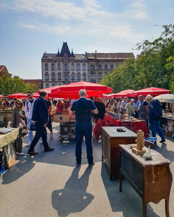 Lots of red umbrellas with antique furniture under it and lots of people around looking at the items at the Britanski Trg Market in Zagreb, Croatia "How to Affordably See Zagreb"