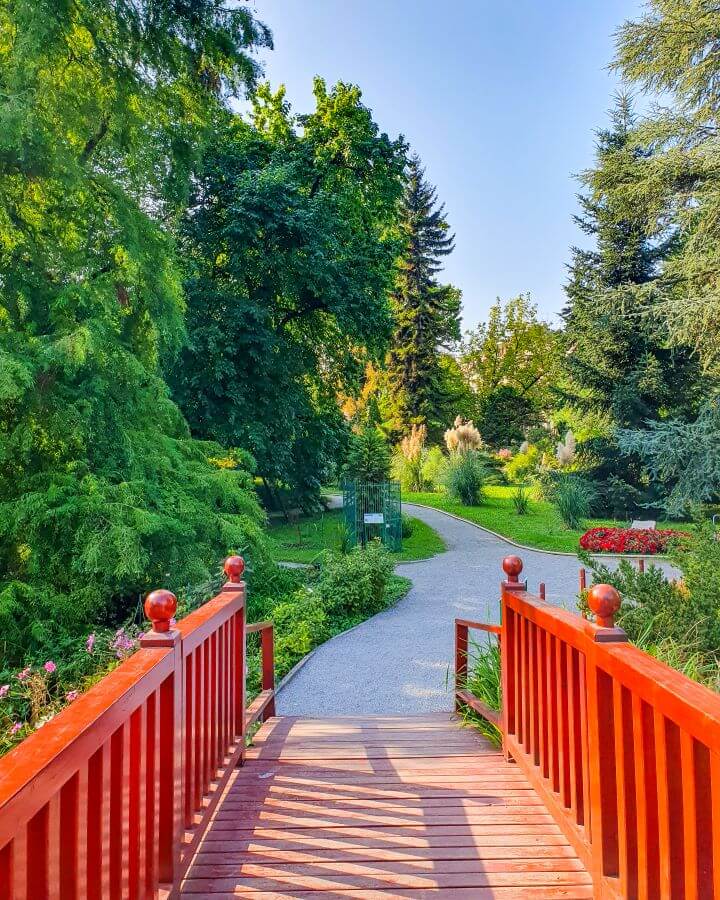 A bright red bridge leading to a rocky path with lots of green grass and trees around the path at the Zagreb Botanic Gardens in Croatia "How to Affordably See Zagreb"