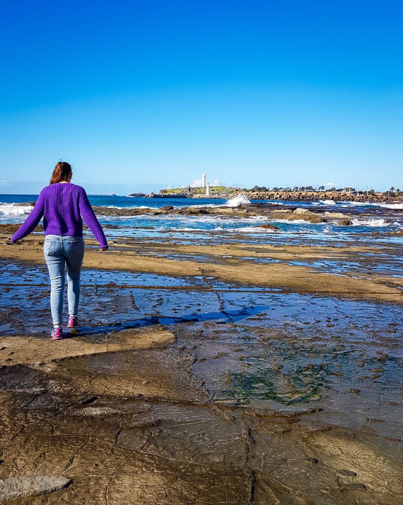 Krissie walking along the edge of the ocean on some rocks. In the distance Flagstaff lighthouse can be seen sitting on a headland in Wollongong, NSW "Four Easy Day Trips From Sydney on the train"