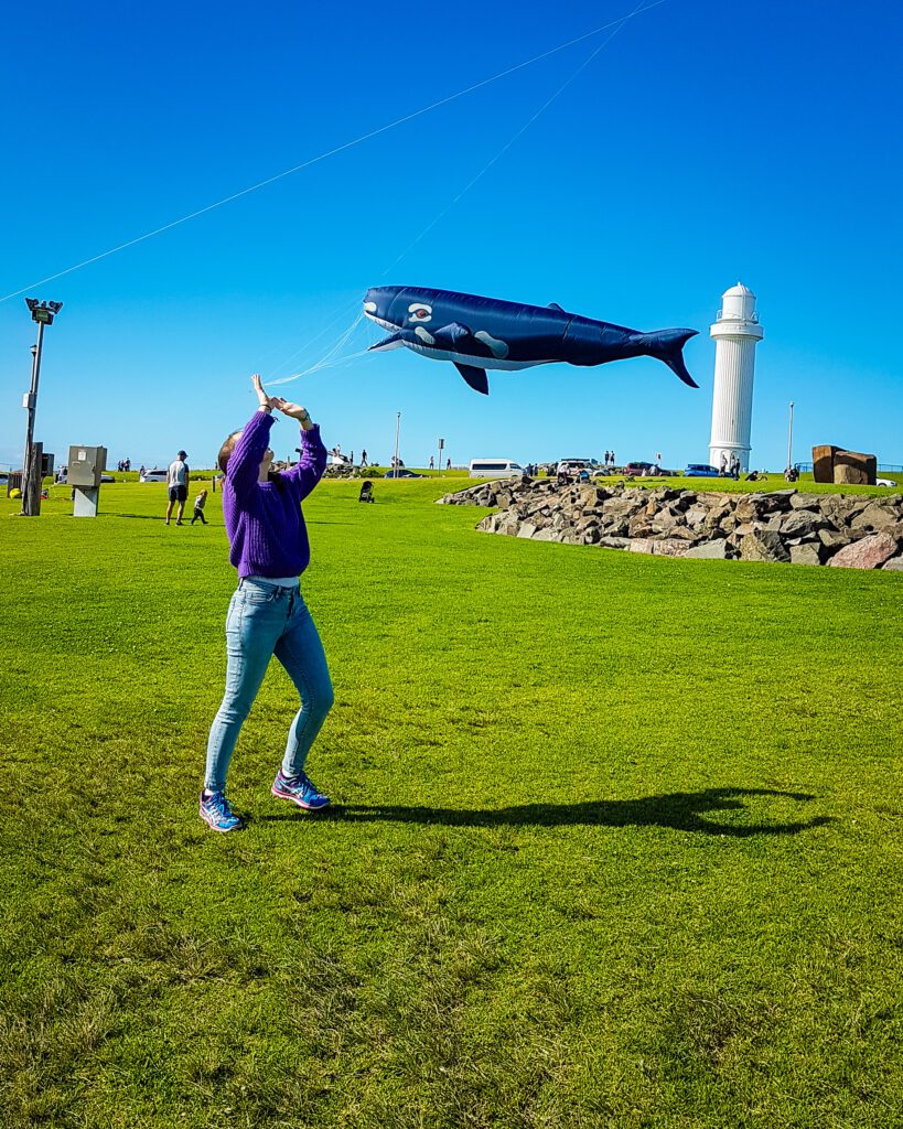 Krissie standing and looking up at a huge kite of a black whale on a big patch of green grass. Behind the kite is the Flagstaff Point Lighthouse in Wollongong, NSW "Four Easy Day Trips From Sydney on the train"