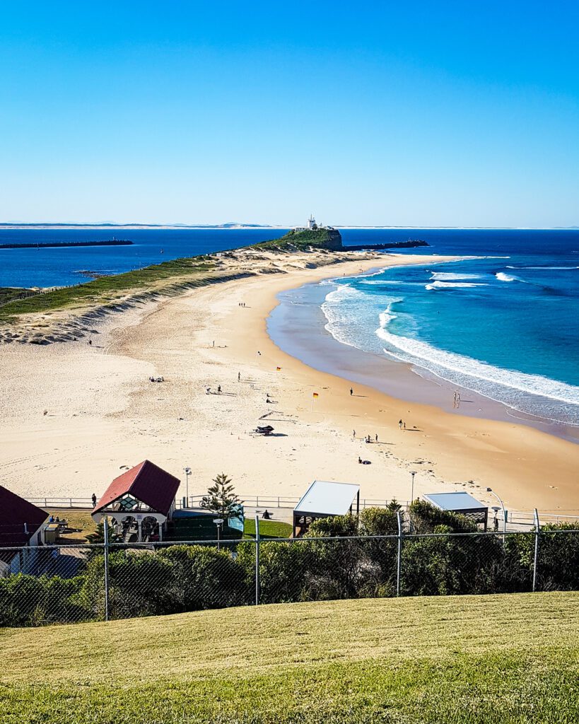 A view down to Nobbys Beach in Newcastle, NSW. Behind the beach is a headland with lots of sand and some green plants and at the end of the headland is Nobbys lighthouse. The photos was taken on a hill so the view is taken from above "Four Easy Day Trips From Sydney on the train"