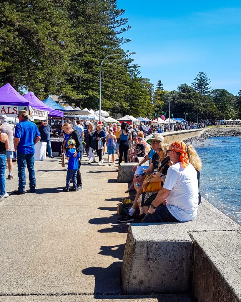 Lots of stalls set up along the water in Kiama, NSW. There are lots of people walking along the stalls looking at them and some people sitting on the seawall "Four Easy Day Trips From Sydney on the train"