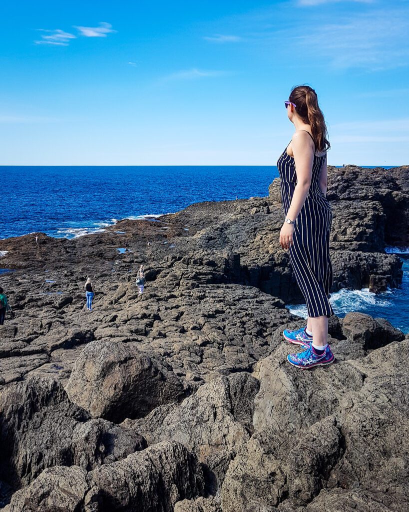 Krissie wearing a black and white striped jumpsuit and standing on some rocks looking out at the ocean in Kiama, NSW. Around her are more brown rocks and the sky and ocean are a brilliant blue colour "Four Easy Day Trips From Sydney on the train"