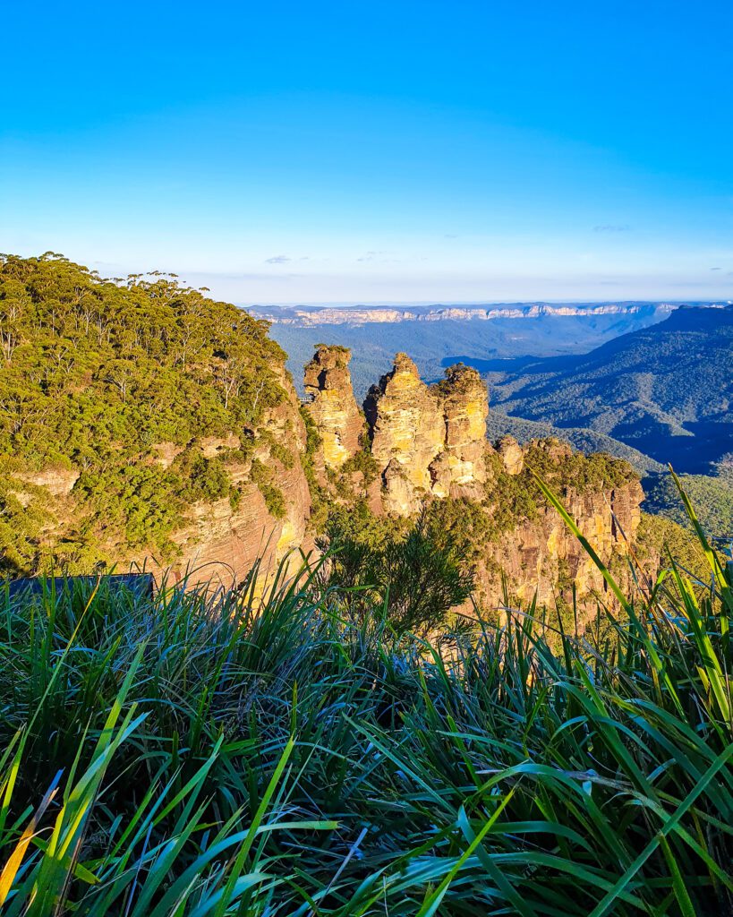 The 3 sisters rock formation in the Blue Mountains, NSW which is 3 sections of rock where the cliff has broken off around them. What's left is 3 huge bits of rock next to each other sticking up on the side of the mountain. Behind the formation is the valley with lots of trees and a blueish tinge and in the foreground is some long green grass and trees "Four Easy Day Trips From Sydney on the train"