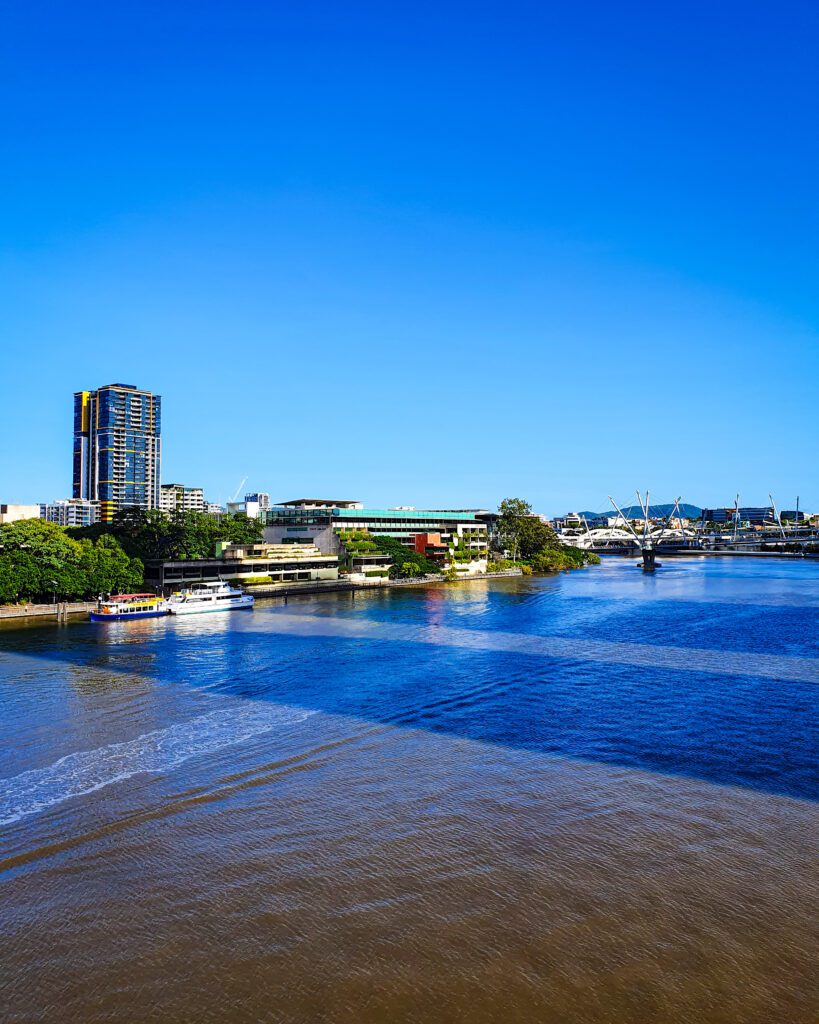 The muddy Brisbane river with some green trees and buildings on the left side of the bank in Brisbane, QLD. Above it is a clear blue sky and another bridge in the distance "How to See Brisbane in a Weekend"