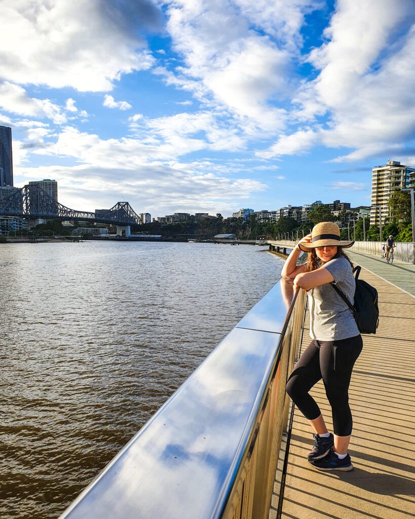 Krissie standing on the New Farm River Walk over Brisbane River in QLD. The walk has a metal handrail and there's a bike passing in the background. You can also see Story Bridge in the distance and above the muddy river is a blue sky with lots of clouds "How to See Brisbane in a Weekend"