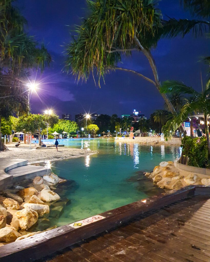 A photo of Streets Beach in Southbank, Brisbane, QLD. The photo is taken at night but there are floodlights that illuminate the sand and blue water. There are also palm trees and rocks around and the whole thing looks super inviting and lovely "How to See Brisbane in a Weekend"