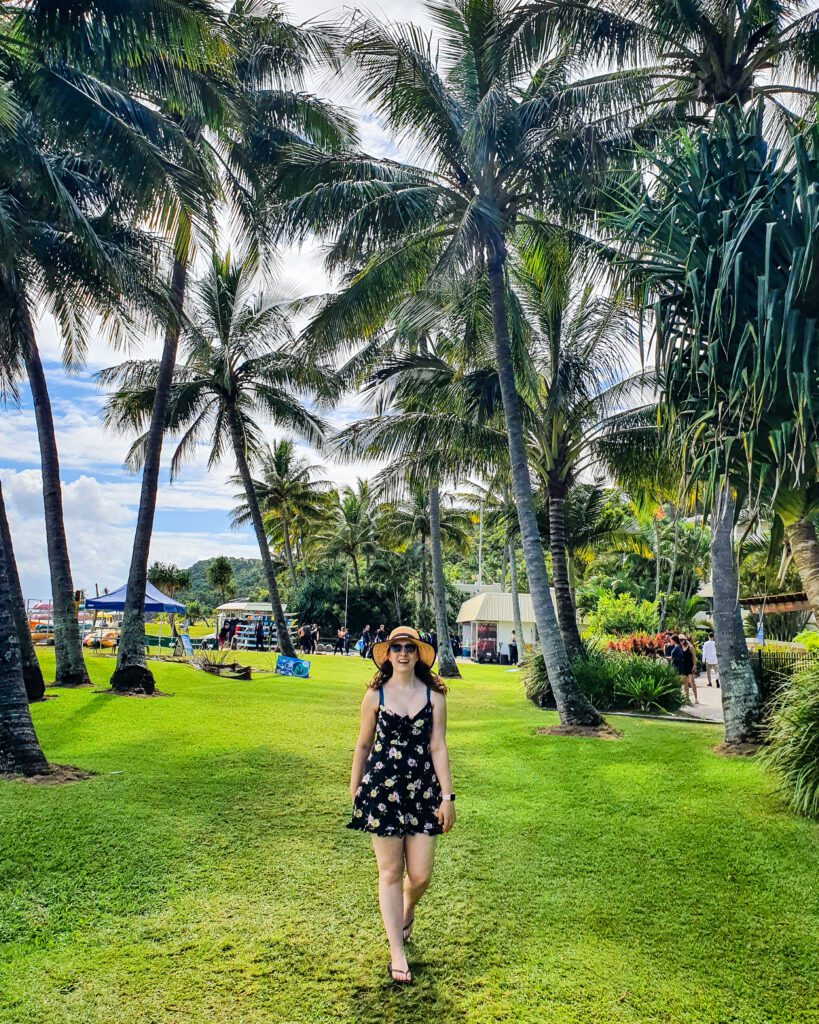 Krissie walking towards the camera on Moreton Island, QLD. There's green grass under her stretching back and lots of palm trees around. In the distance is a tent with people around hiring snorkelling gear "How to See Brisbane in a Weekend"