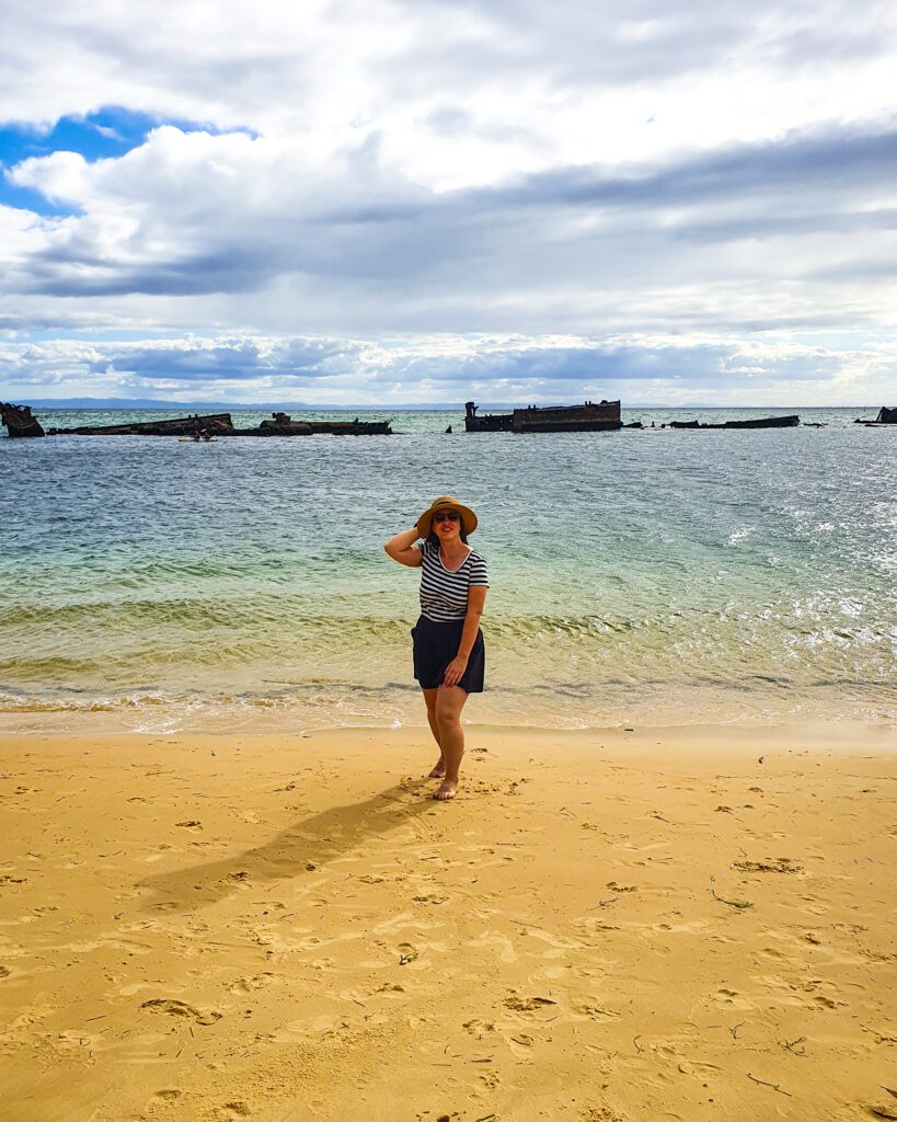 Katherine standing on a beach on Moreton Island, QLD. She's standing on a sandy beach with the ocean behind her and out in the water are the remains of a shipwreck "How to See Brisbane in a Weekend"