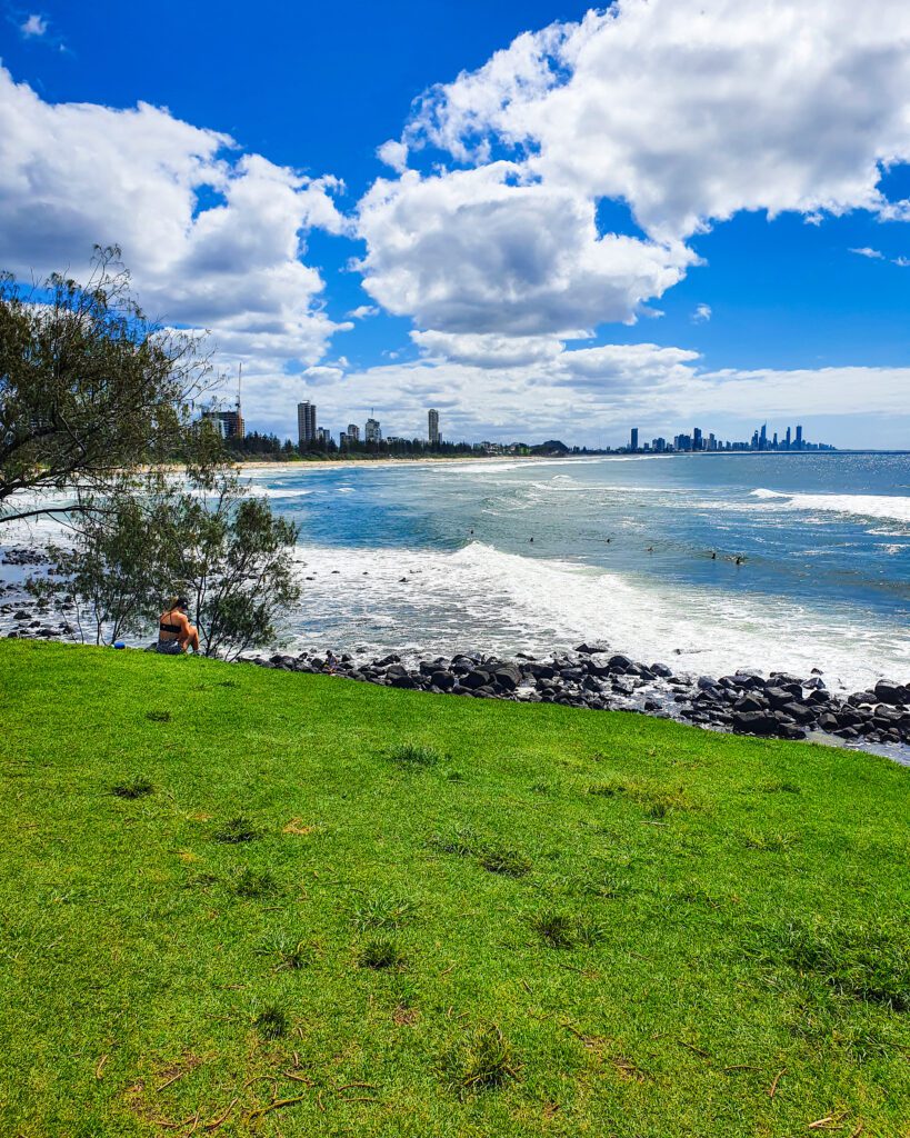 The foreground of the photo is lots of green grass and beyond that are some black rocks and the blue ocean with lots of white waves. Beyond that you can see lots of high rise buildings in Surfers Paradise on the Gold Coast in QLD "How to See Brisbane in a Weekend"