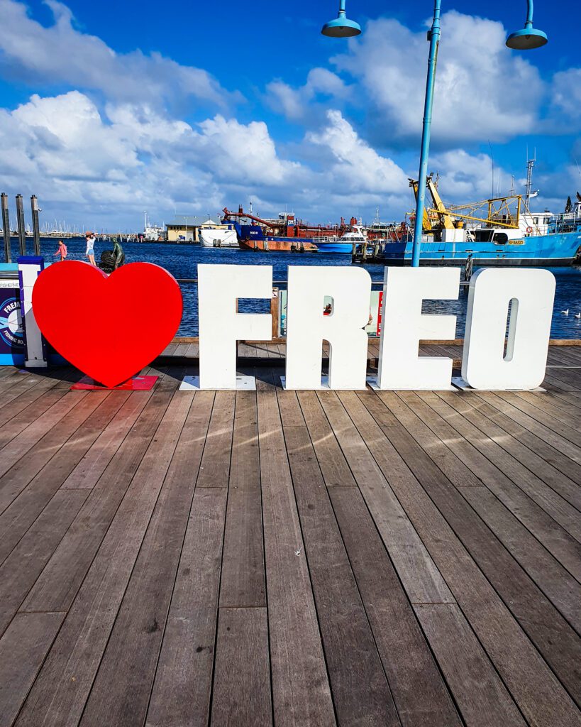 A big sign with white letters that say "I" and "Freo" with a big red love heart in between in Fremantle, WA. In front of the sign is some wooden flooring and behind it is the water, some fishing boats and the blue sky "How to See Perth in a Weekend"