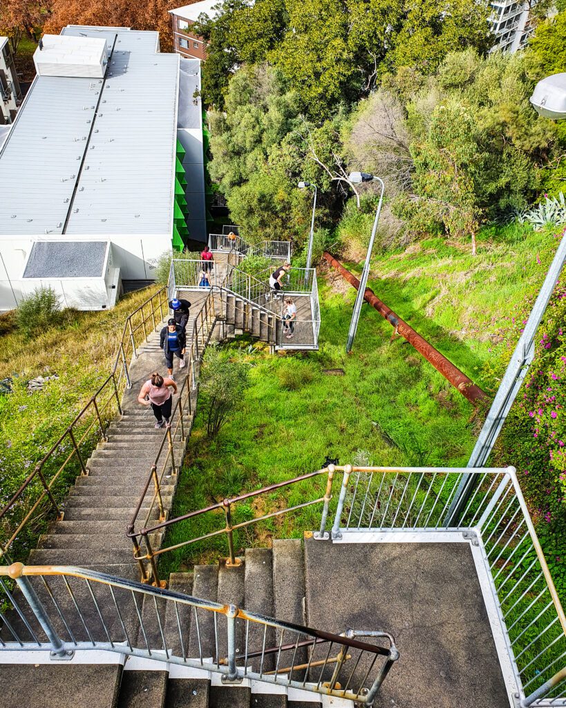 A steep concrete staircase in Perth, WA with metal railings taken from above with people running and walking up it. Around the staircase is green grass and trees and the roof of a building "How to See Perth in a Weekend"