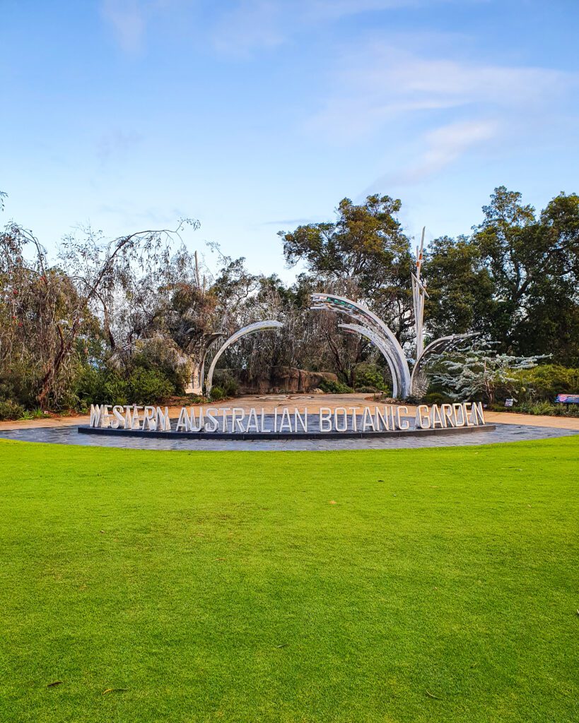 A large patch of short neat green grass with a metal sign on the ground that says Western Australian Botanic Garden in Perth, WA. Above it are some trees and the blue sky "How to See Perth in a Weekend"