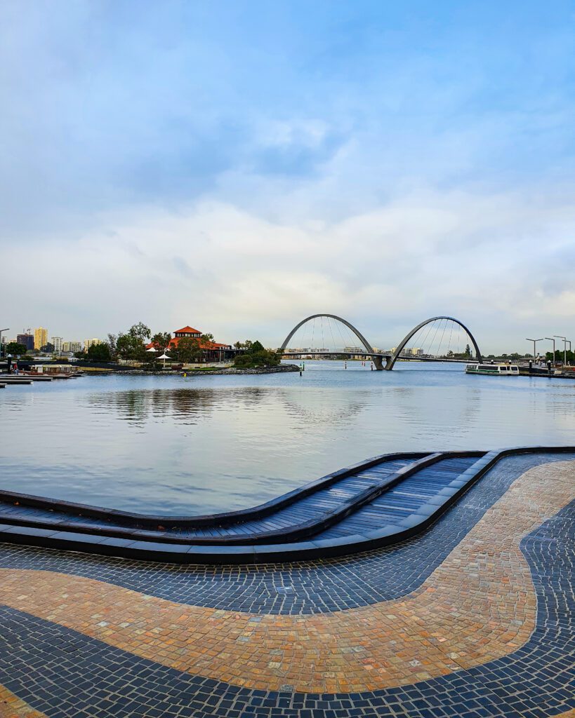 A tiled walkway in the foreground with a bridge with 2 arches in the background leading from the mainland to the island in Elizabeth Quay, Perth WA. On the island you can see trees and a building with an orange roof "How to See Perth in a Weekend"