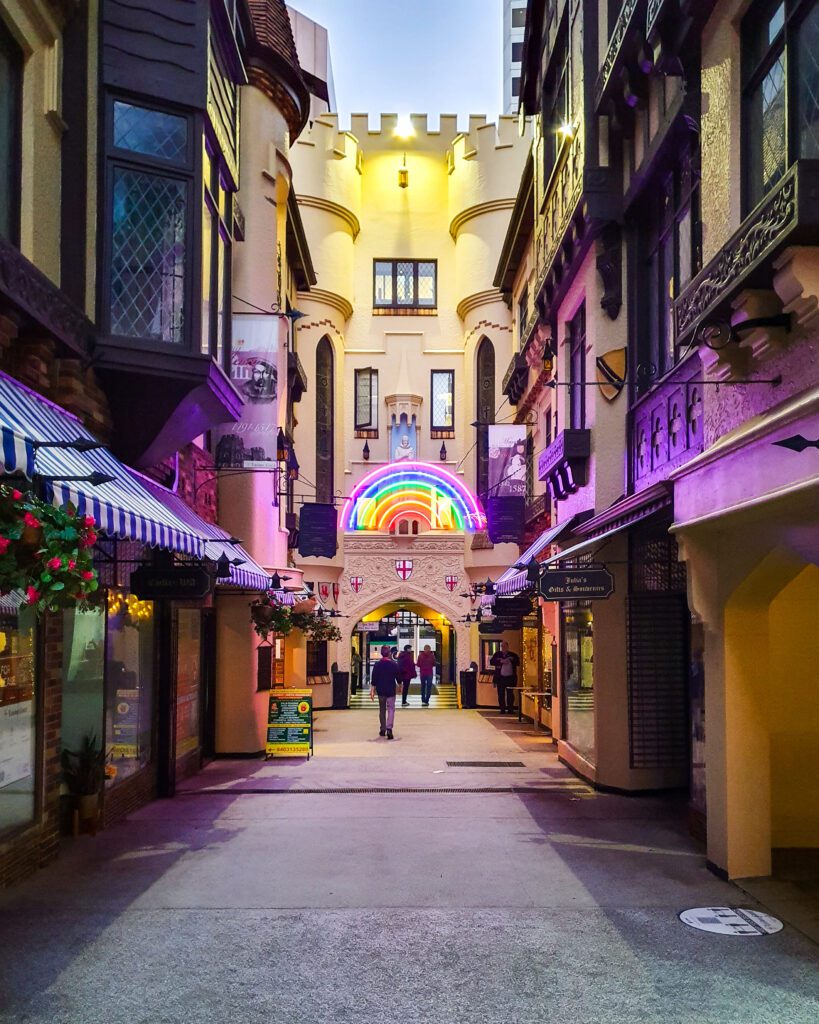 A narrow street in Perth city with Tudor style architecture with a building at the end of the street that looks like a castle wall. There's a light up rainbow sign on the wall at the end and not a lot of people on the street "How to See Perth in a Weekend"