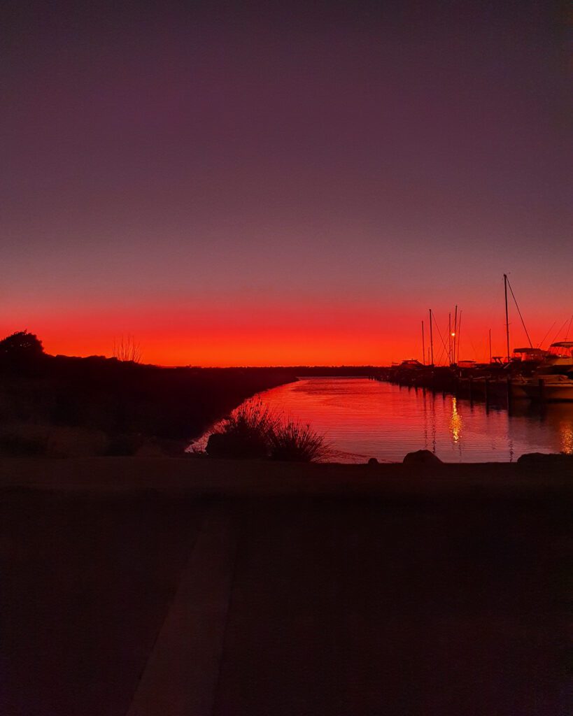 How to See Perth in a Weekend. A photo of an incredible deep red and orange sunset that fades up into dark grey and black. The sunset is behind a boat harbour so is reflecting in the water and some boats and plant silhouettes can be seen in the foreground in Fremantle, WA