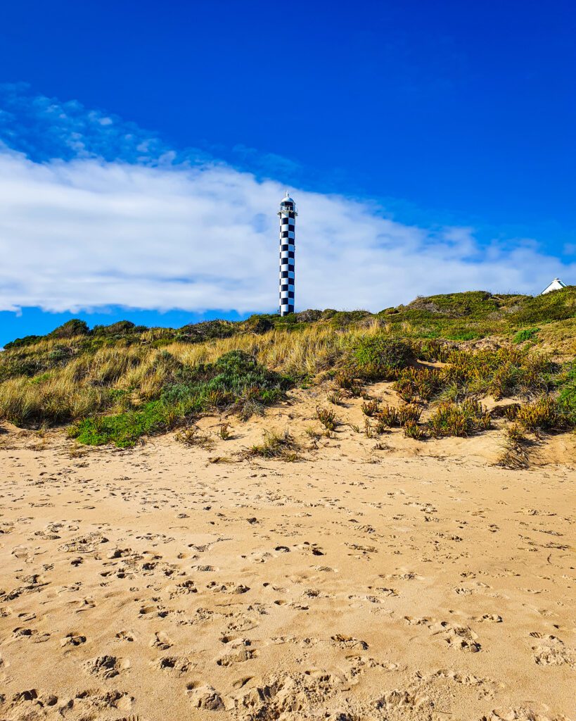 A photo of a beach in Bunbury, WA with lots of footprints on it. Behind the sand is a hill with some green and yellow grass and a tall skinny black and white checked lighthouse in the middle "How to See Perth in a Weekend"