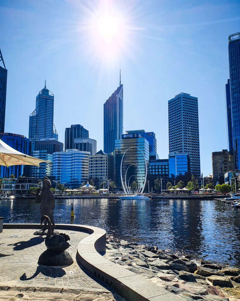 A view back to Perth city and its high rise buildings from the island in Elizabeth Quay, Perth WA. In front of the high rise buildings is a white sculpture of 1 large oval with 4 smaller ovals inside it. On this side of the island is a metal sculpture of a woman and a swan and the sun is shining "How to See Perth in a Weekend"