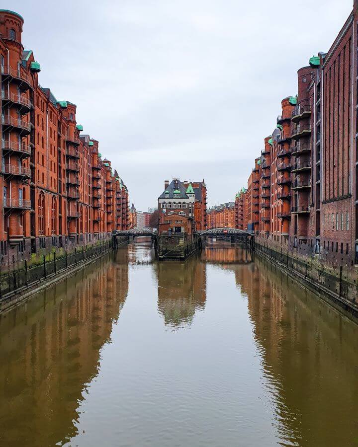 The famous view in Speicherstadt in Hamburg, Germany. The view is of a big warehouse area with red buildings around a river and two bridges leading to an island in the middle of the river "How to See Hamburg on a Budget"